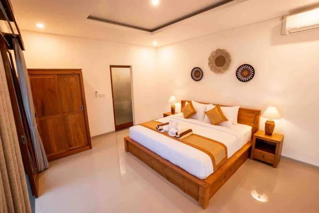 A well-lit modern bedroom featuring a large wooden bed with white and gold bedding, two nightstands with lamps, and a wooden wardrobe.