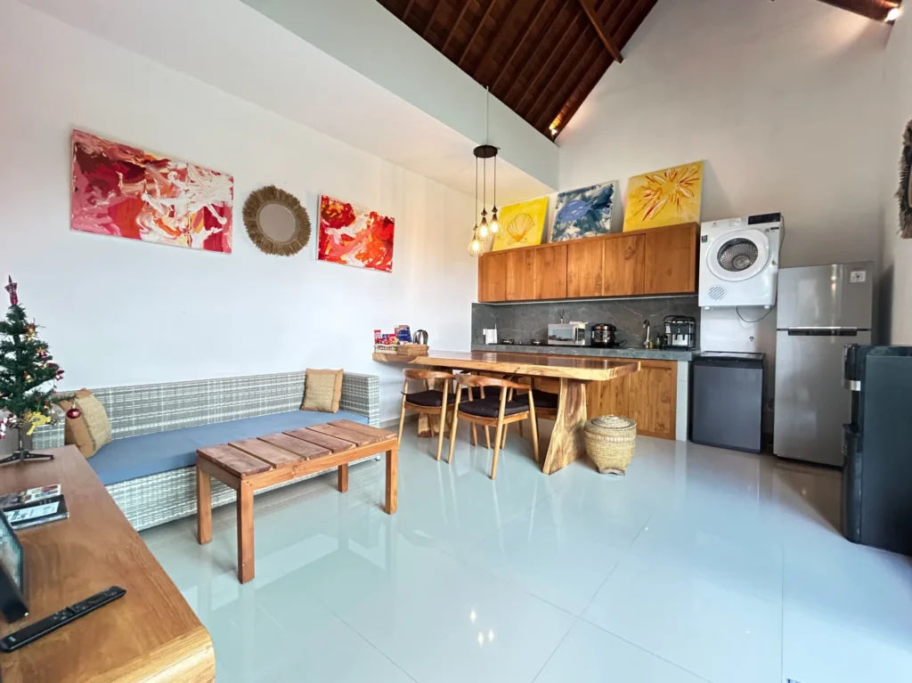 Bright open-plan living room with modern kitchen, dining area, sofa, coffee table, and colorful abstract art. Features high ceilings and a small Christmas tree.