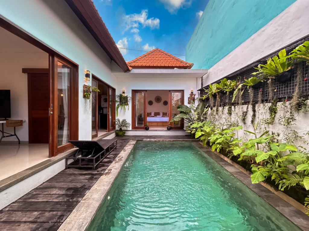 A modern Balinese-style villa with a long, narrow private swimming pool, wooden deck, lush plants, and open glass doors revealing interior rooms under a blue sky.
